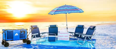 A beach setup with a cart labeled "OCEANTRAX" and a sign indicating "Complementary Beach Gear" including chairs, towels, cooler, cart, umbrella, and blanket.