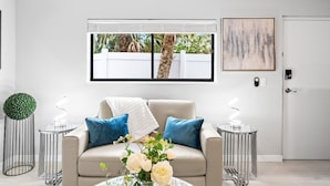 A modern living room with a beige sofa, two blue pillows, a white throw, and a vase with white flowers. There are two metal side tables with white lamps. A large window with blinds shows a white fence and trees outside. A framed abstract painting hangs on the wall above the door. - Beach Rd, 300ft to Beach Access & 500ft to Siesta Key Village, 1bdr (Siesta Key)