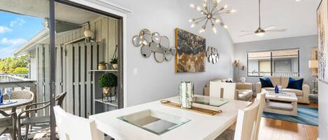 A modern living room with a glass dining table, white sofas, and a ceiling fan.