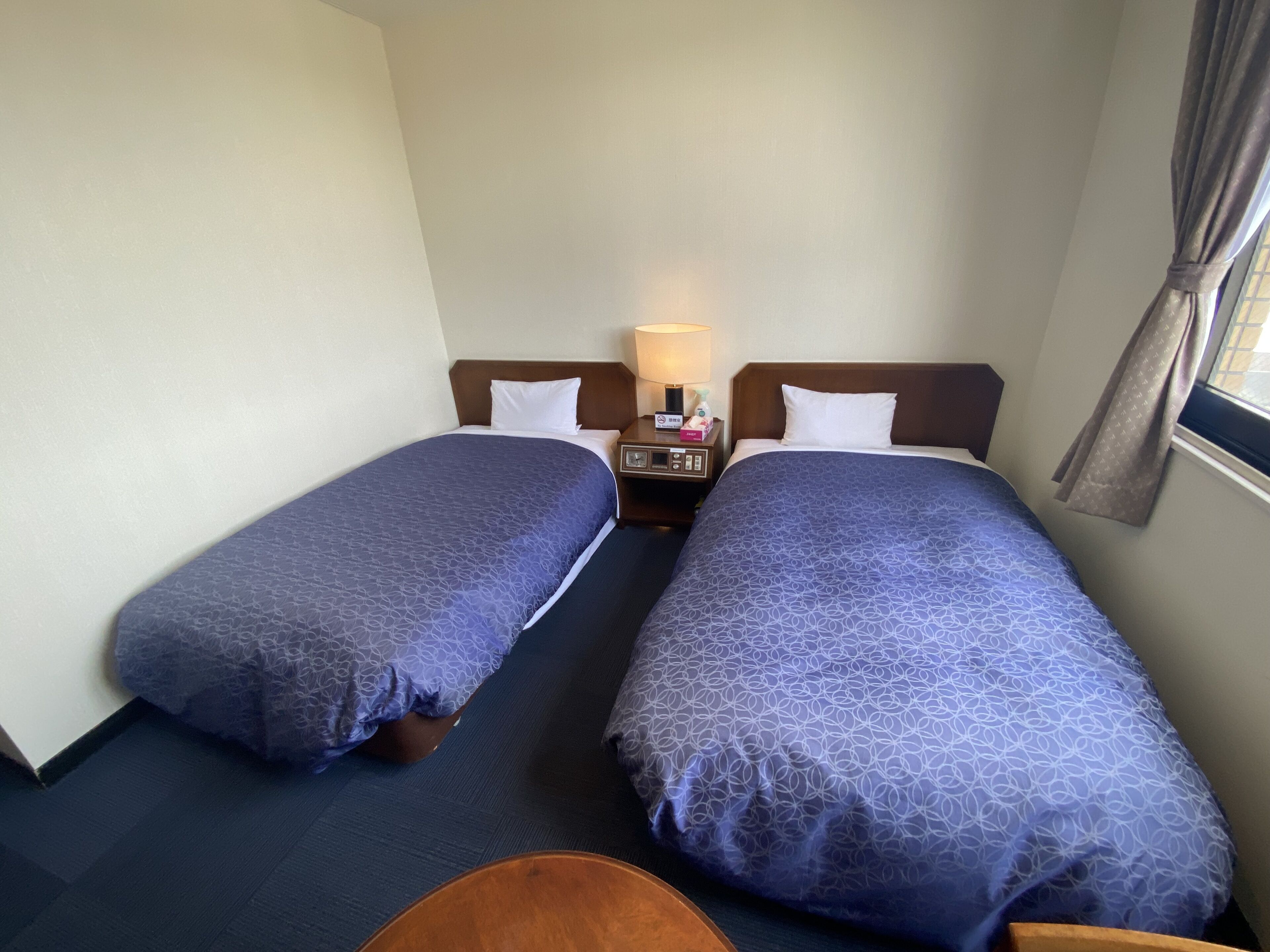 A hotel room with two single beds, a nightstand with a lamp, a small table, and a window with curtains.
