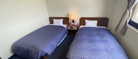 A hotel room with two single beds, a nightstand with a lamp, a small table, and a window with curtains.