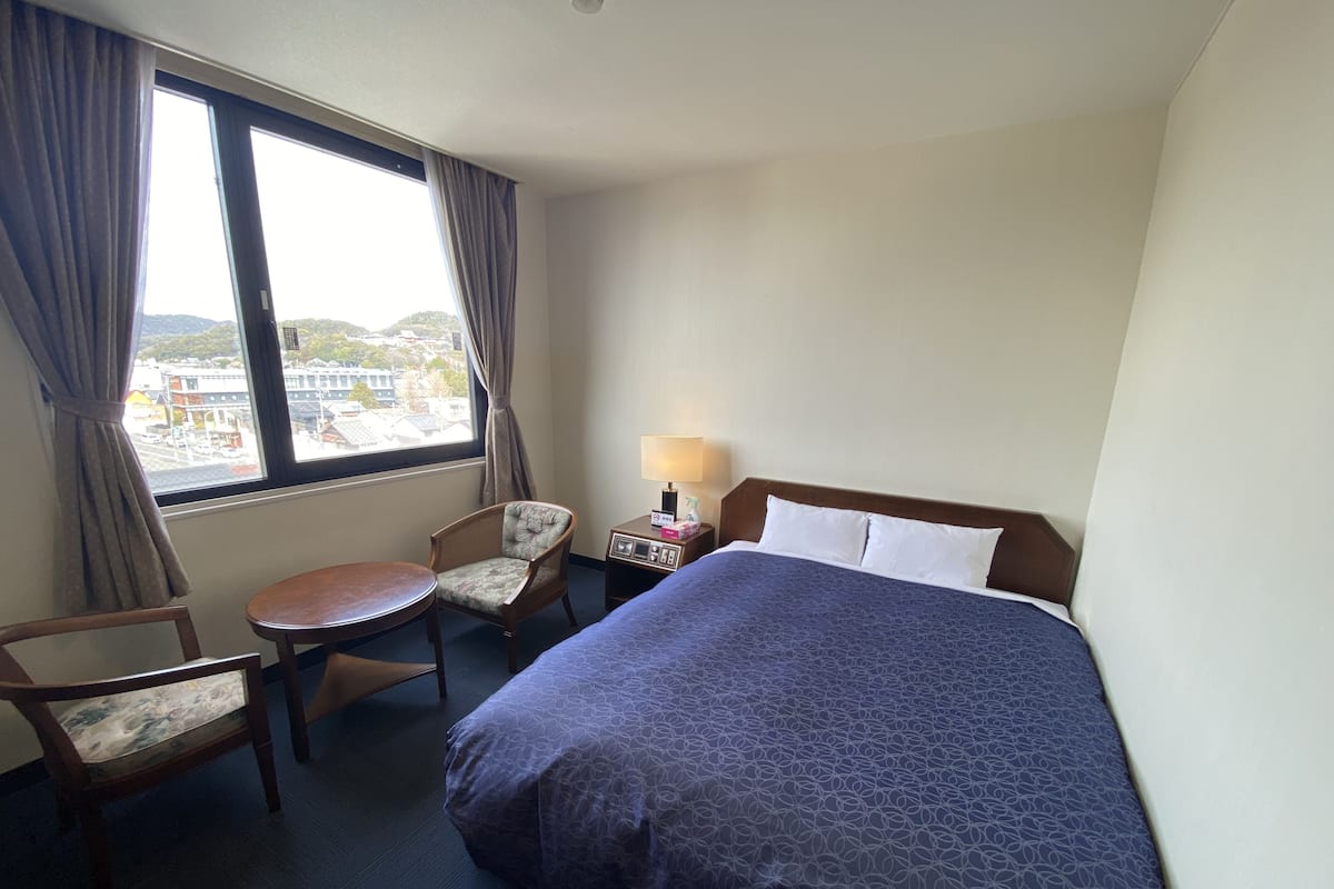 Une chambre d’hôtel avec un lit, une chaise, une petite table, une table de chevet et une fenêtre avec des rideaux.
