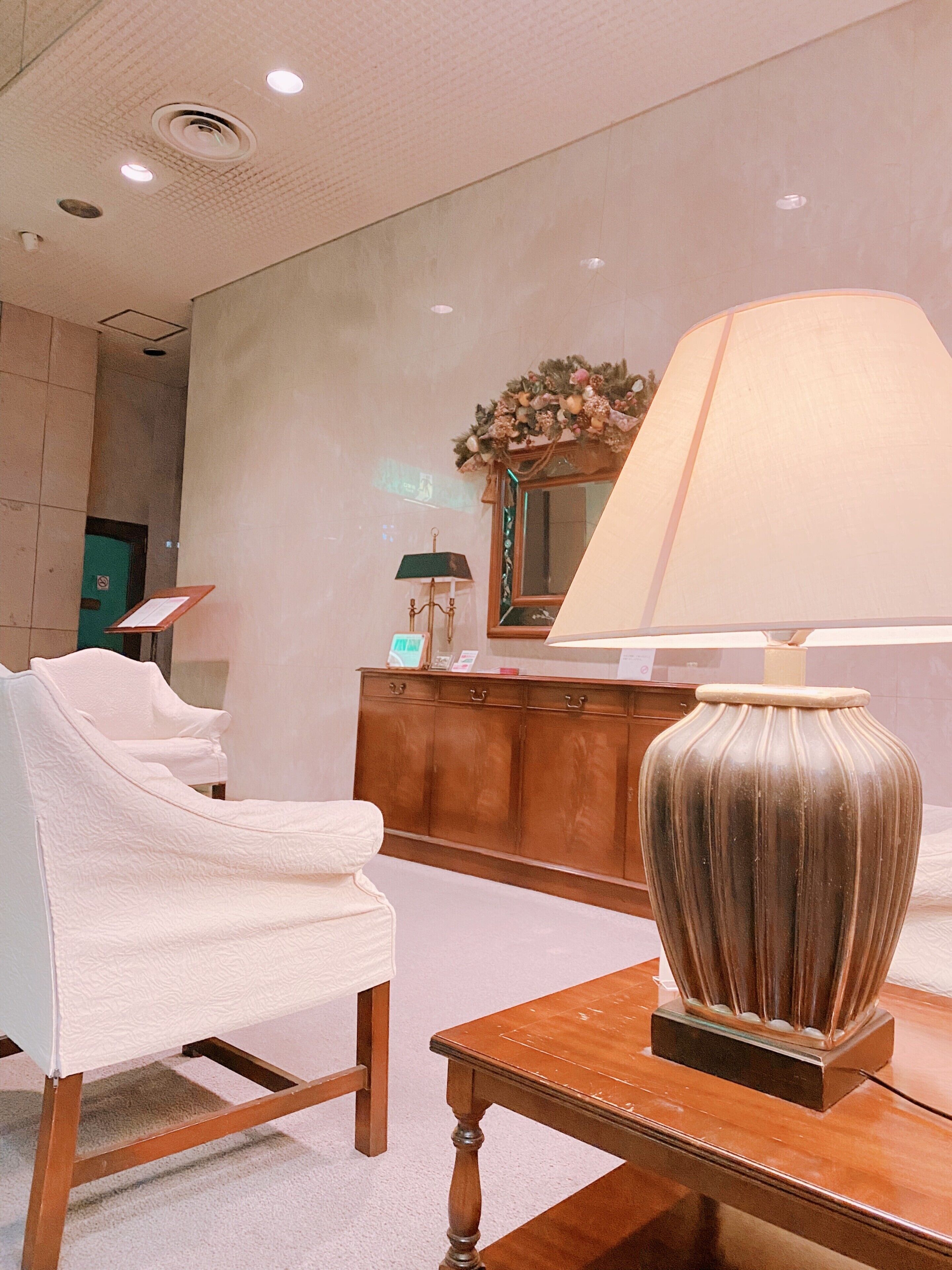 A hotel room with a white armchair, a wooden table with a lamp, and a mirror with a floral decoration.