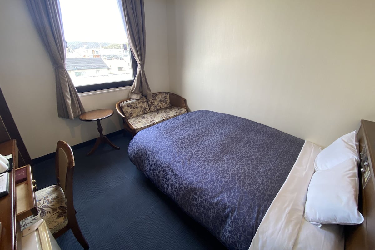 Une chambre d’hôtel avec un lit, une chaise, un bureau et une fenêtre avec des rideaux.