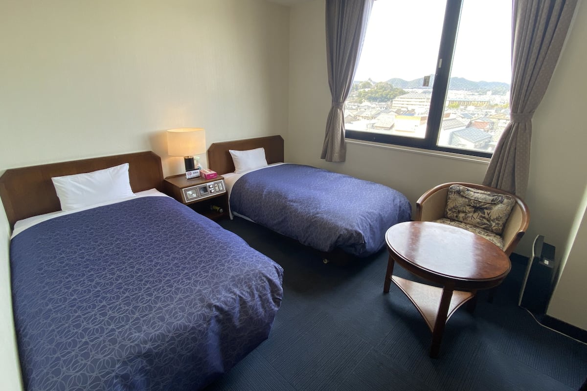 Une chambre d’hôtel avec deux lits, une table de chevet avec une lampe, une chaise et une fenêtre donnant sur des immeubles.