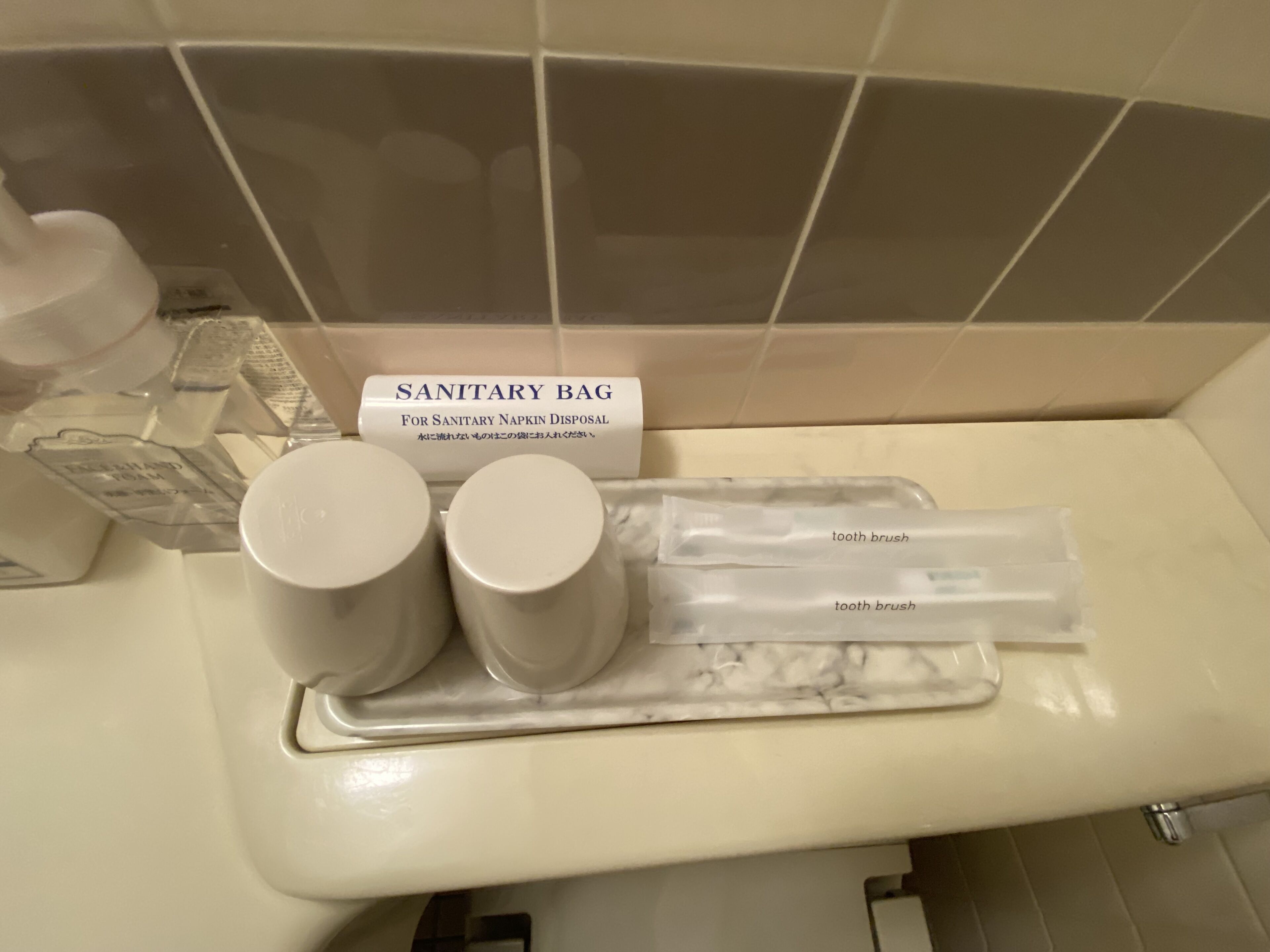 A bathroom sink area with a sanitary bag dispenser, two cups, and two toothbrushes.
