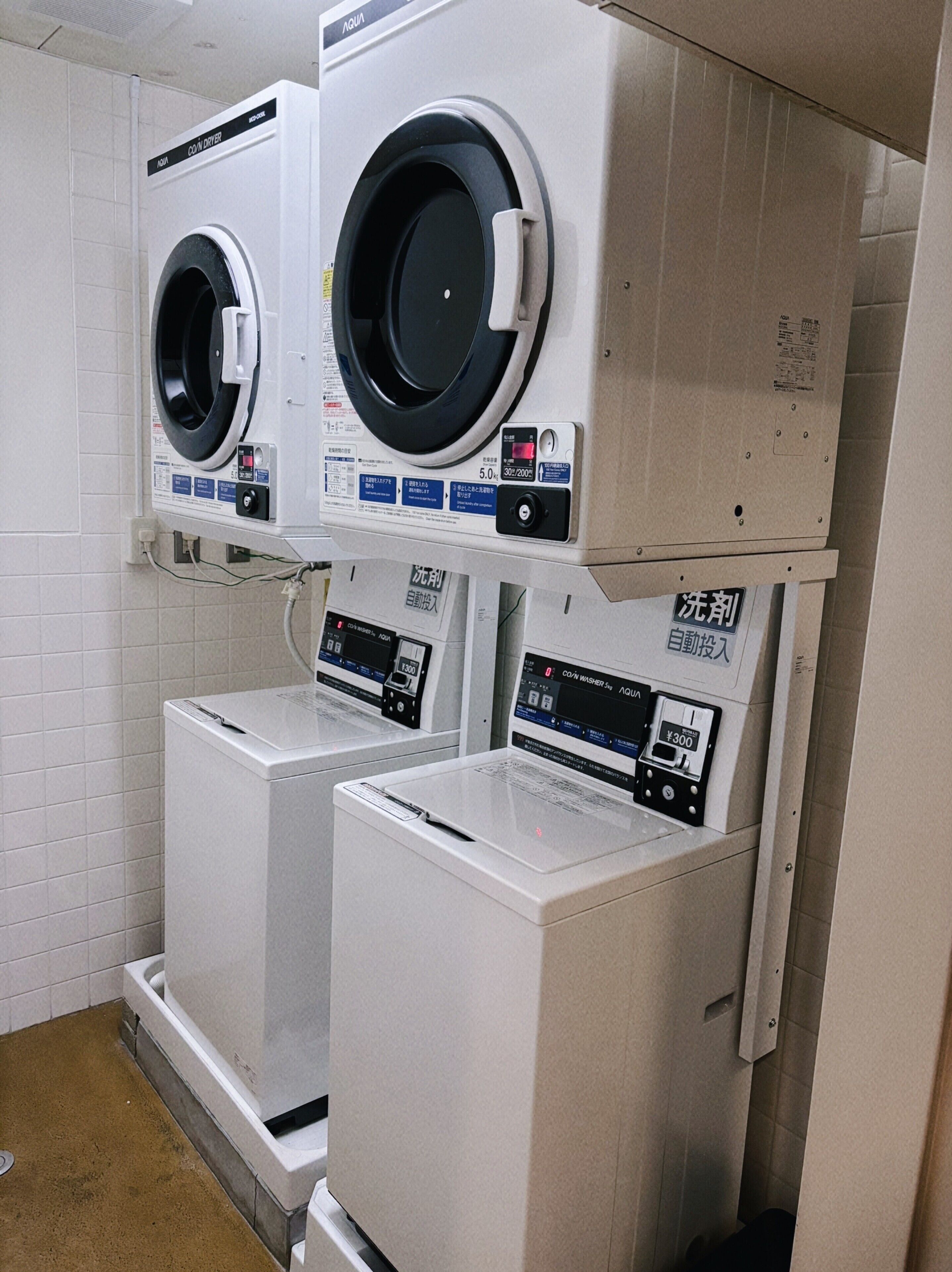 A laundry facility with two coin-operated washers and dryers, control panels, and a tiled floor.