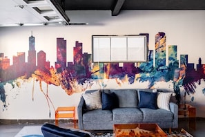 Una habitación con un mural de un paisaje urbano colorido, un sofá gris y una ventana con persianas.