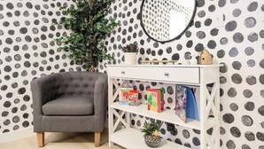 Une pièce avec un fauteuil gris, une console blanche ornée de livres et d’une petite plante, et un papier peint à pois.