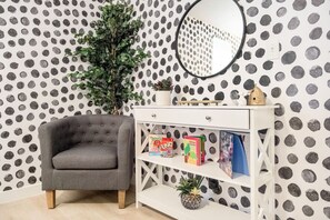 Una habitación con un sillón gris, una consola blanca con libros y una pequeña planta, y un papel pintado con lunares.