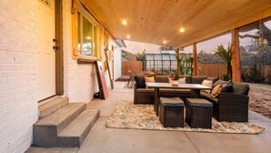 Ein überdachter Patio-Bereich mit Gartenmöbeln, einem großen Fenster und einer Holzbalkendecke.