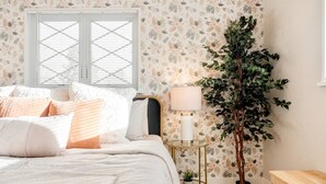 A bedroom with a bed, a nightstand with a lamp, a potted plant, and a window with blinds.