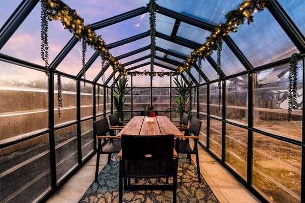 A greenhouse-style room with a wooden table and chairs, large windows, and hanging plants.