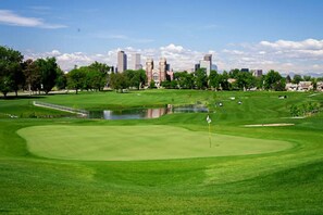A golf course with a water hazard, a flagstick, and a city skyline in the background.