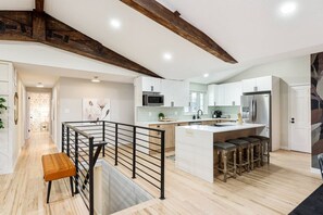 Cocina moderna con armarios blancos, una isla central con taburetes, una escalera de madera y vistas al área de estar.