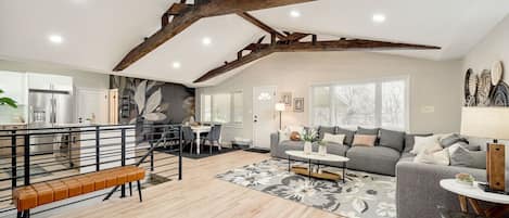 A spacious living area with a high ceiling, exposed wooden beams, a grey sofa, a dining table, and a kitchen in the background.