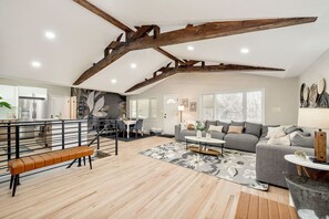 A spacious living area with a high ceiling, exposed wooden beams, a grey sofa, a dining table, and a kitchen in the background.