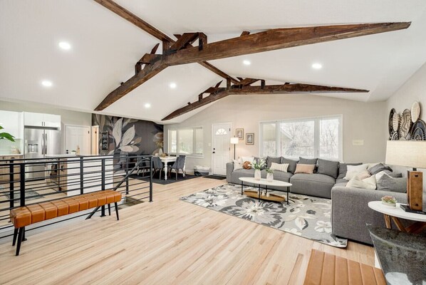 A spacious living area with a high ceiling, exposed wooden beams, a grey sofa, a dining table, and a kitchen in the background.
