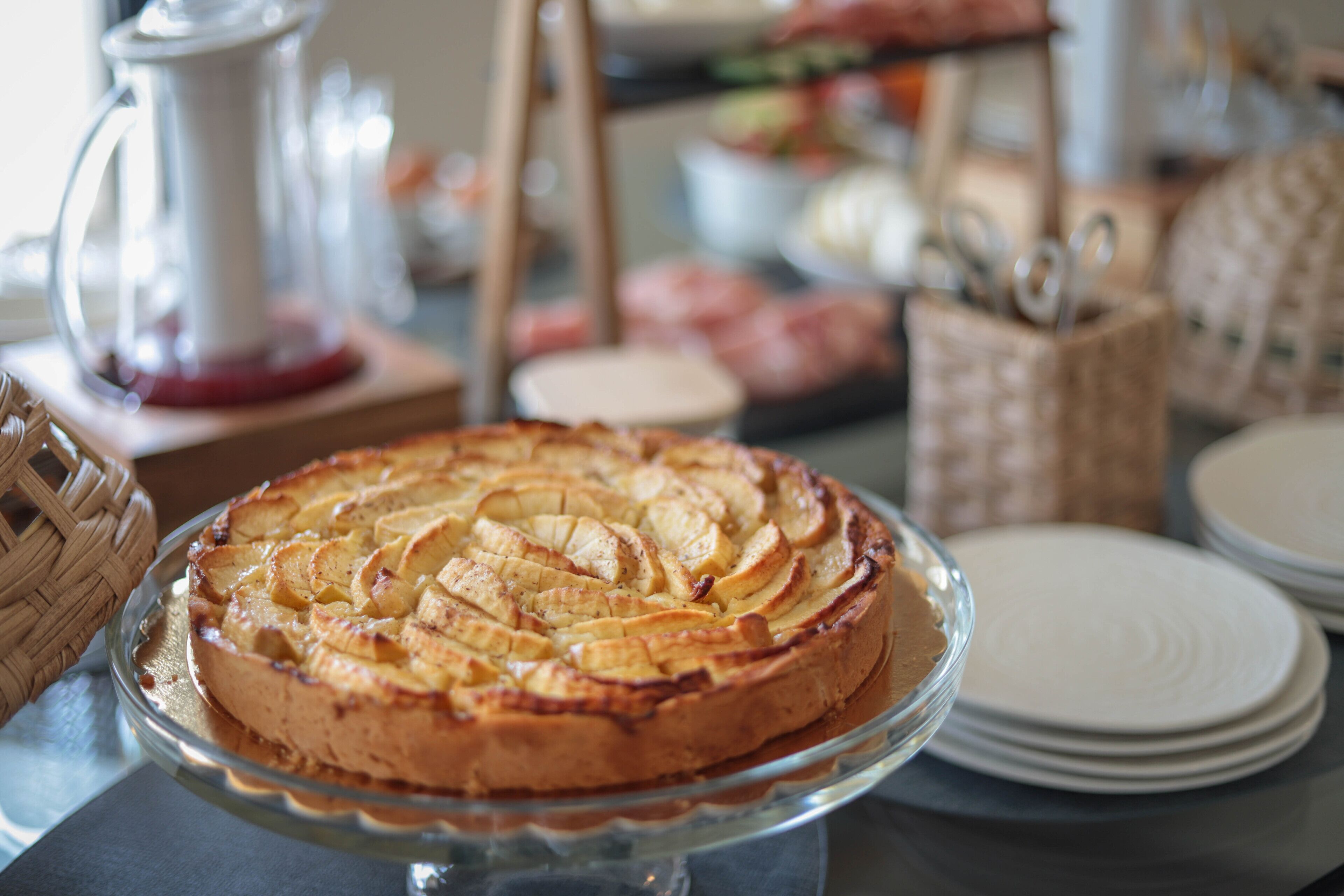Un pastel redondo con rodajas de manzana sobre un pedestal de vidrio, rodeado de platos y cestas.
