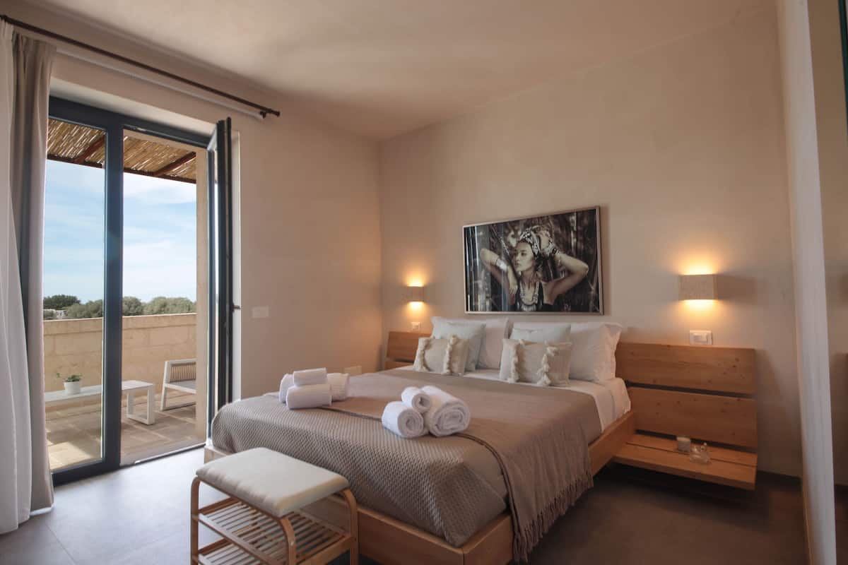 Une chambre à coucher comprenant un lit, des tables de chevet, un banc et une vue sur l’extérieur, accessible par une porte coulissante.