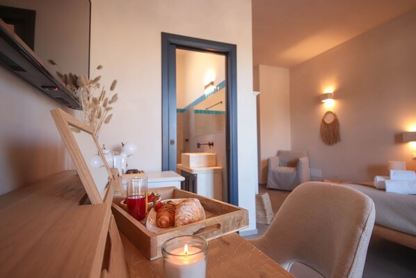 Une pièce chaleureuse avec une table en bois, une bougie, un plateau de pâtisseries et une boisson, et un lit en arrière-plan.