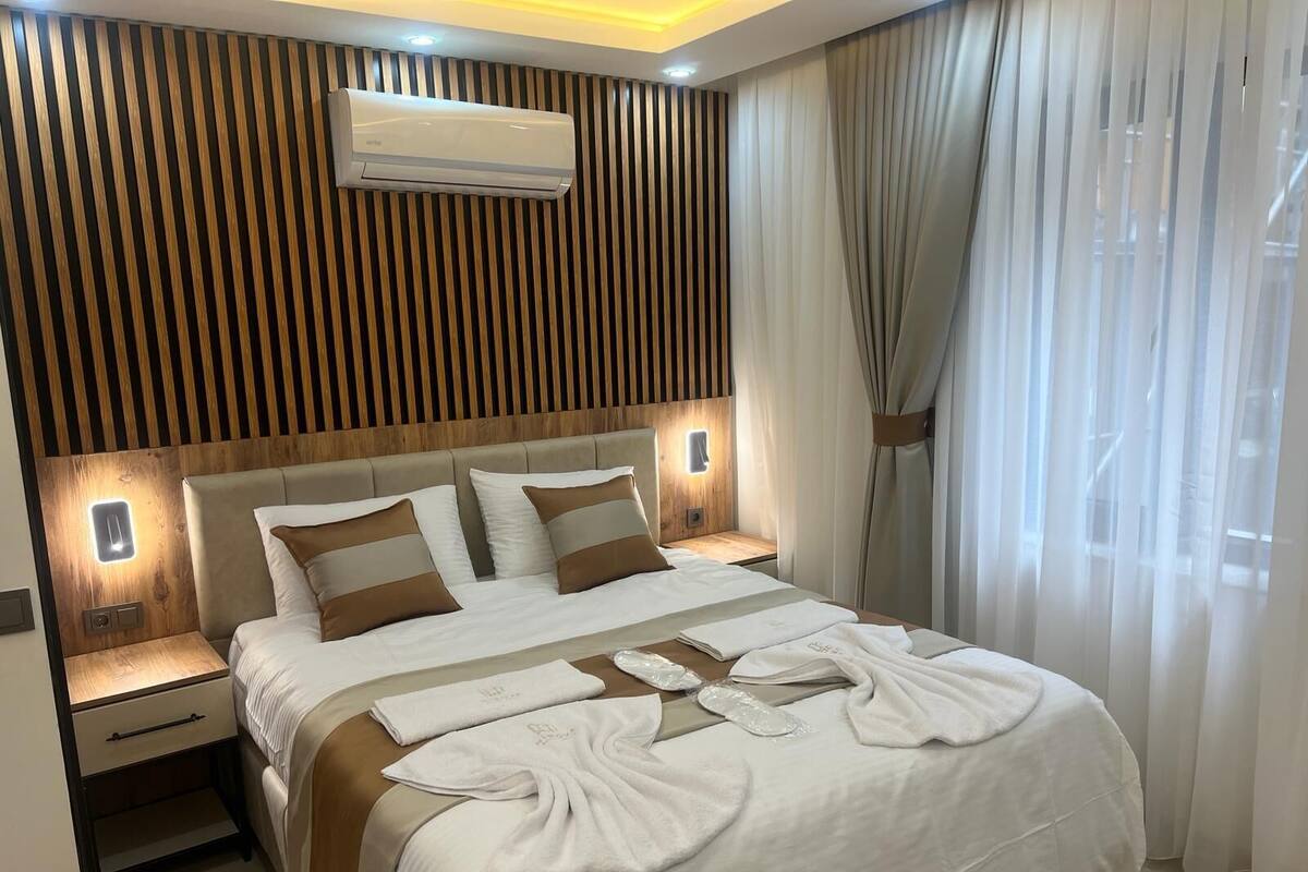 A modern hotel room with a wooden headboard, a large bed with white and brown bedding, a bedside table with a lamp, and a window with sheer curtains.