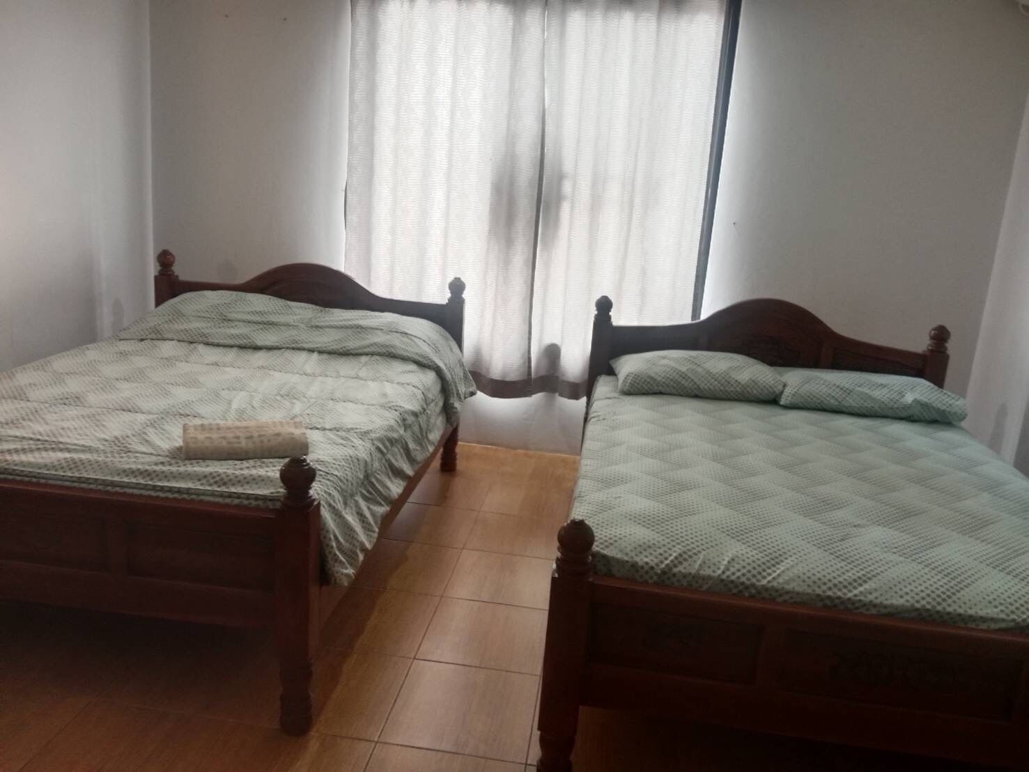 Une chambre avec deux lits en bois, chacun recouvert d’une courtepointe matelassée et agrémenté d’oreillers. La chambre a un sol carrelé et une fenêtre ornée de rideaux légers.