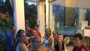Eine Gruppe von Menschen sitzt an einem Holztisch in einer Außenumgebung mit einer Glasscheibe und einem Ventilator.