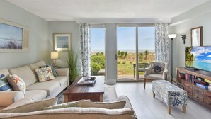A living room with a sofa, coffee table, and TV stand, large sliding glass doors leading to a view of the outdoors. - Villas By The Sea #184 - Oceanfront (Jekyll Island)