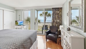 A bedroom with a bed, a TV, a chair, a dresser, and a balcony with a view of the sea. - Villas By The Sea #184 - Oceanfront (Jekyll Island)