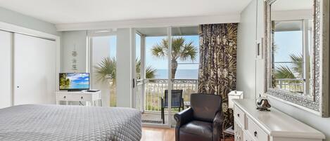 Een slaapkamer met een bed, een televisie, een stoel, een dressoir en een balkon met uitzicht op zee.