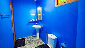 A bathroom with blue walls, a white sink, a toilet, and a window with a view of greenery. - Sunbird's Home  (Udawalawa)