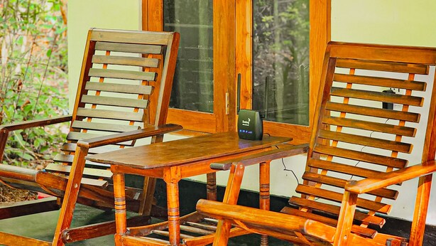 A wooden rocking chair and a wooden chair with a table in front of a window with a view of greenery. - Sunbird's Home  (Udawalawa)