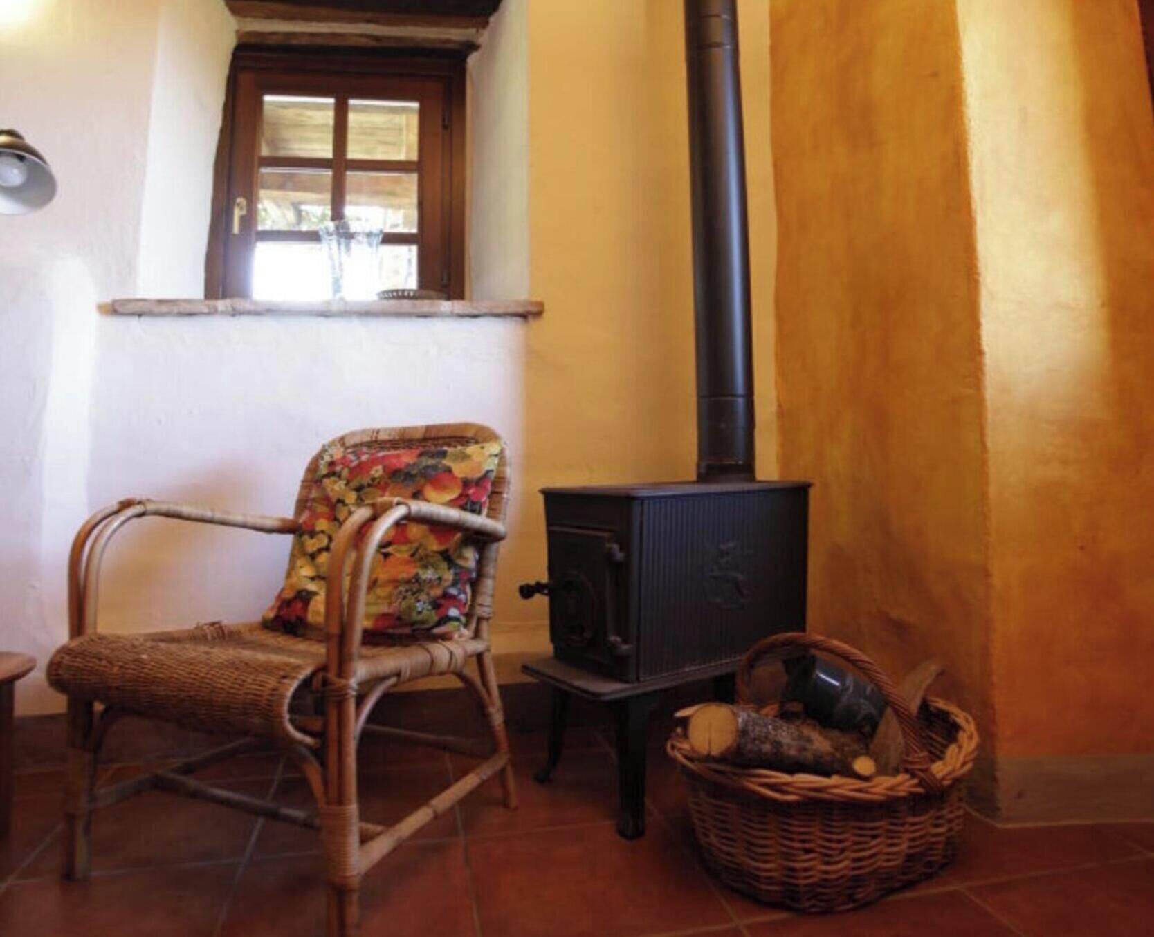 Una habitación rústica con una silla de mimbre, una pequeña mesa de madera, una estufa negra y una cesta de mimbre con leña.