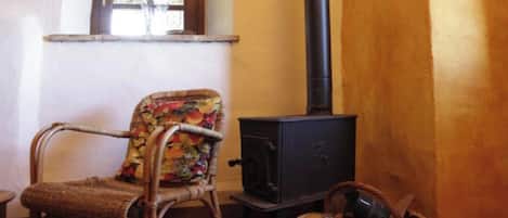 Una habitación rústica con una silla de mimbre, una pequeña mesa de madera, una estufa negra y una cesta de mimbre con leña.