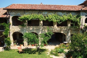 Un edificio de piedra con techo de tejas, ventanas arqueadas y un patio con macetas y una banca.