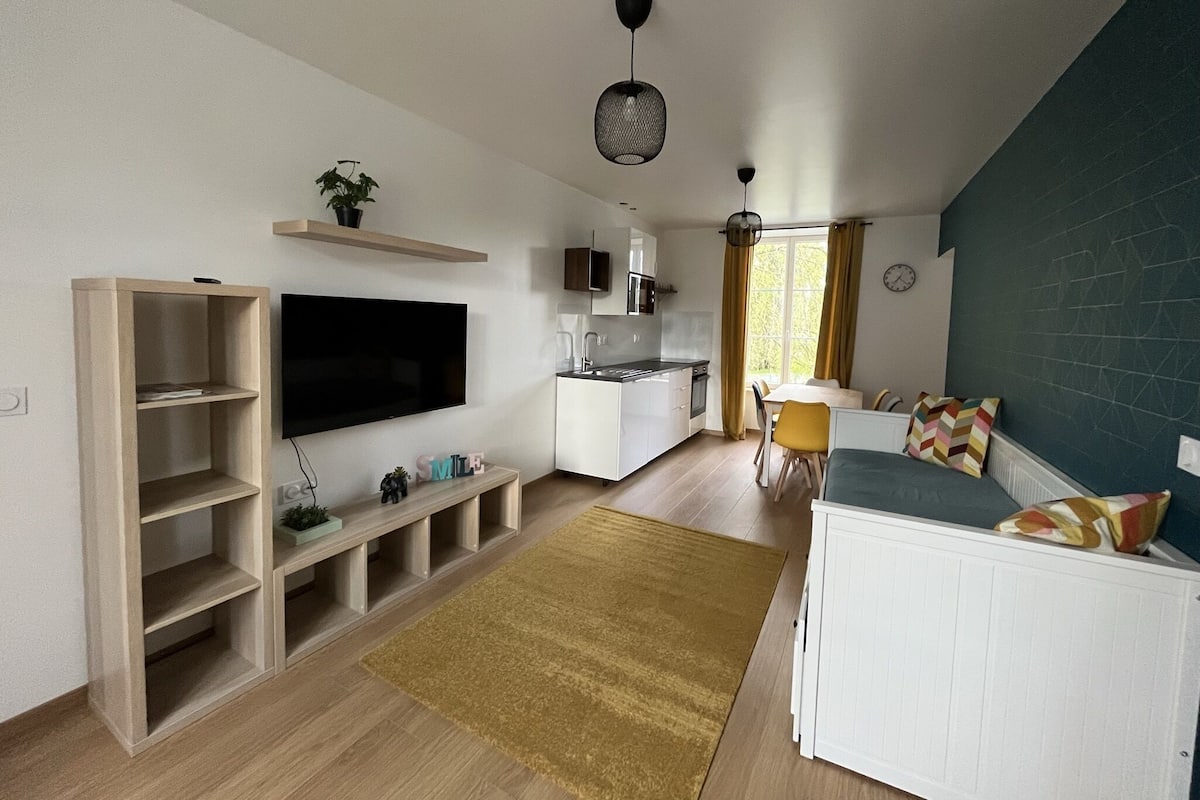 A modern living room with a flat-screen TV, wooden shelving, a sofa, and a kitchen area with white cabinets and a dining table.