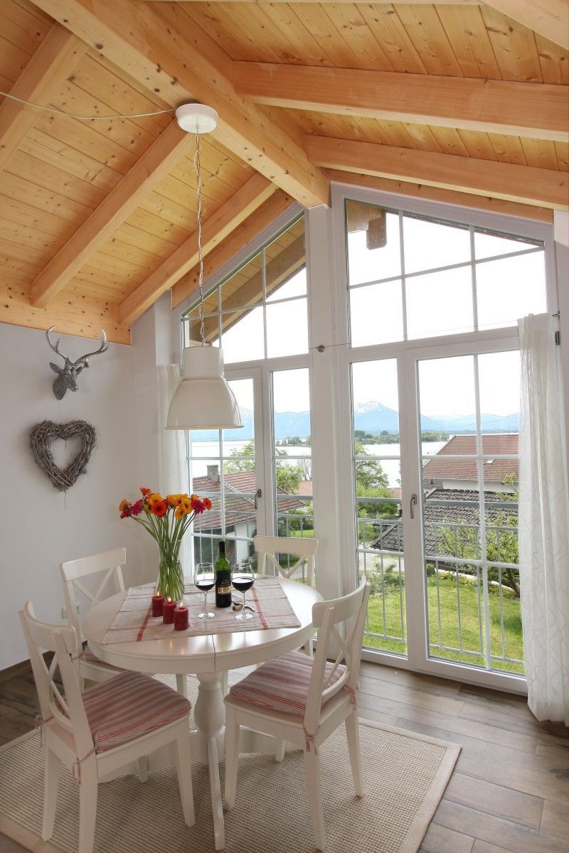 Un coin repas avec une table ronde, des chaises blanches et une vue sur un paysage montagneux, visible à travers de grandes fenêtres.
