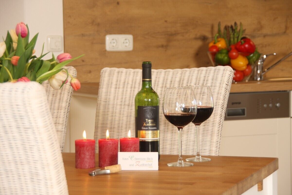 Una mesa de comedor con una botella de vino, dos copas de vino tinto y dos velas encendidas. Se observa un jarrón con tulipanes y un cuenco con verduras coloridas al fondo.