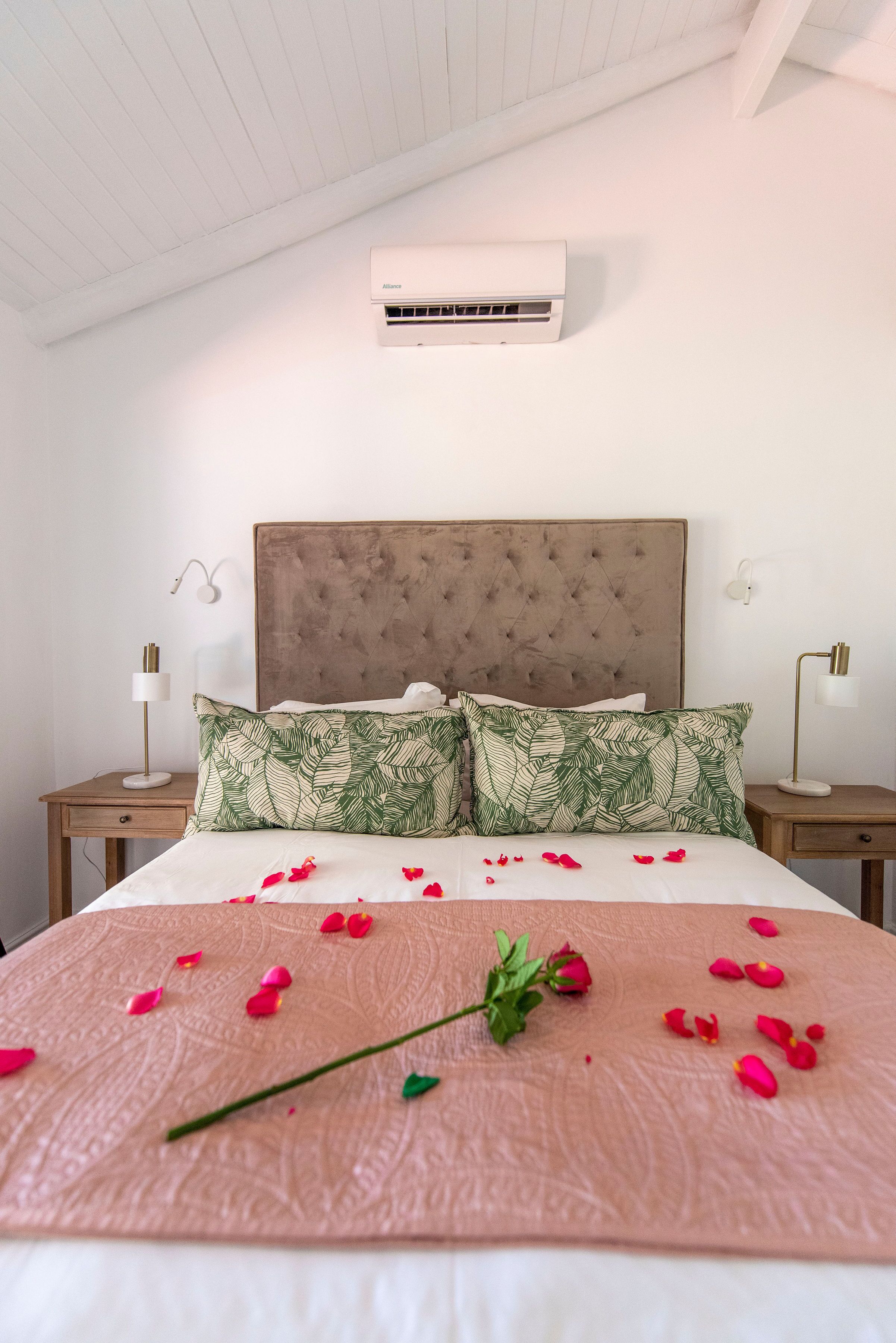 A neatly made bed with a pink textured bedspread, two green leaf-patterned pillows, and scattered rose petals.
