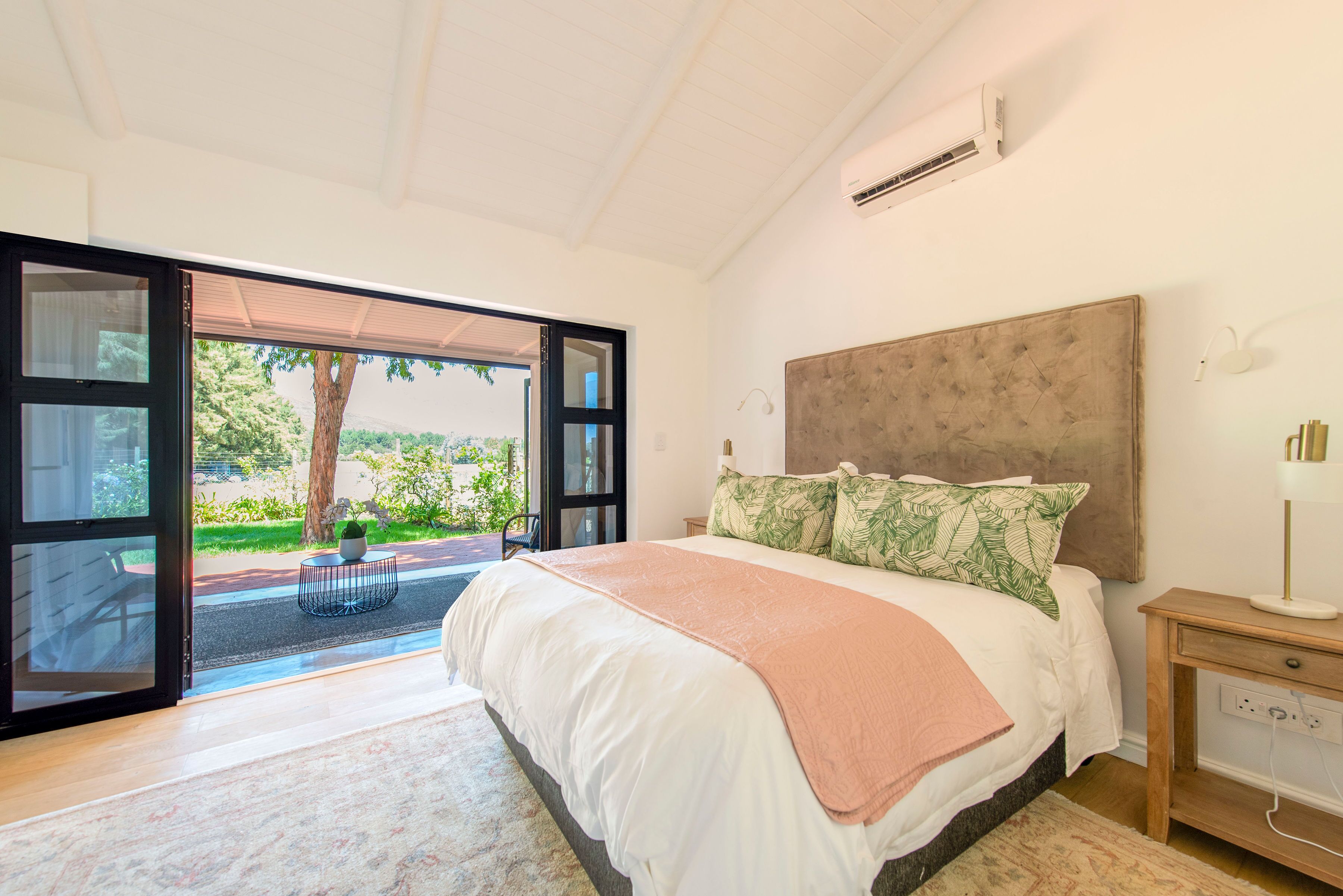 A bedroom with a large bed, a nightstand, and a view through an open door to a garden.