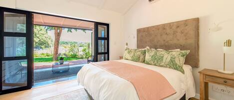 A bedroom with a large bed, a nightstand, and a view through an open door to a garden.