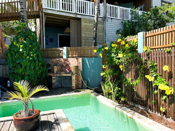 A backyard with a swimming pool, a wooden deck, a brick barbecue area, and a wooden fence.