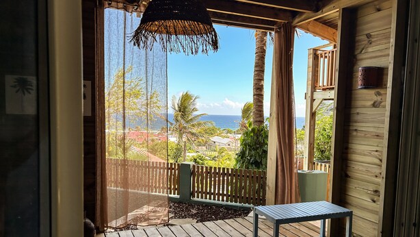 A wooden deck with a table and bench, overlooking a coastal area with palm trees and houses. - Plein la vue (Saint-Pierre)