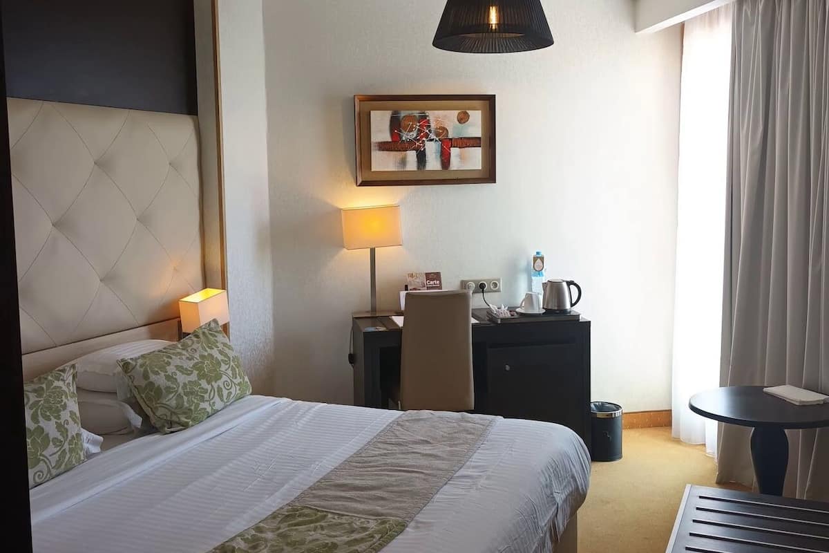 A hotel room with a large bed, a desk with a lamp, a chair, a small table, and a painting on the wall.
