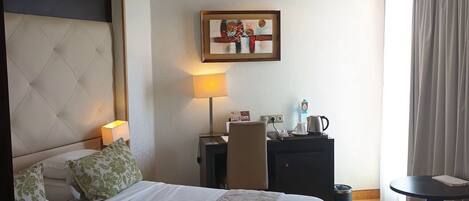 A hotel room with a large bed, a desk with a lamp, a chair, a small table, and a painting on the wall.