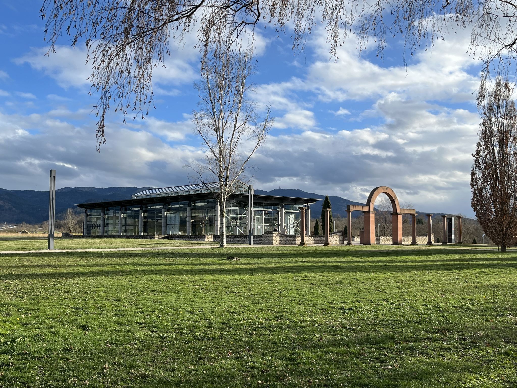 Un edificio moderno con fachada de vidrio, un gran arco y un campo cubierto de césped al frente.