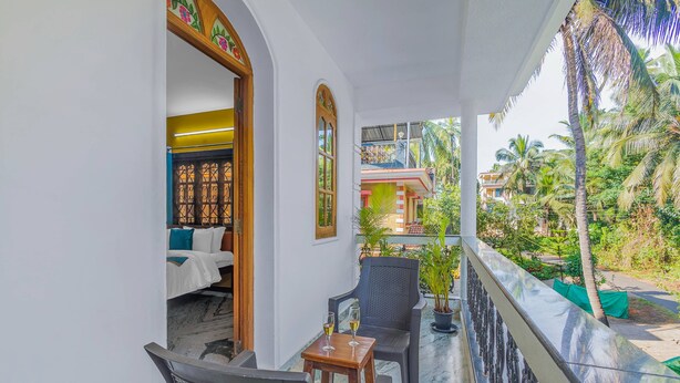 A balcony with a view of a tropical garden, a bed with white bedding, and a small table with two glasses of wine. - The Sequeira Guest House (Majorda)