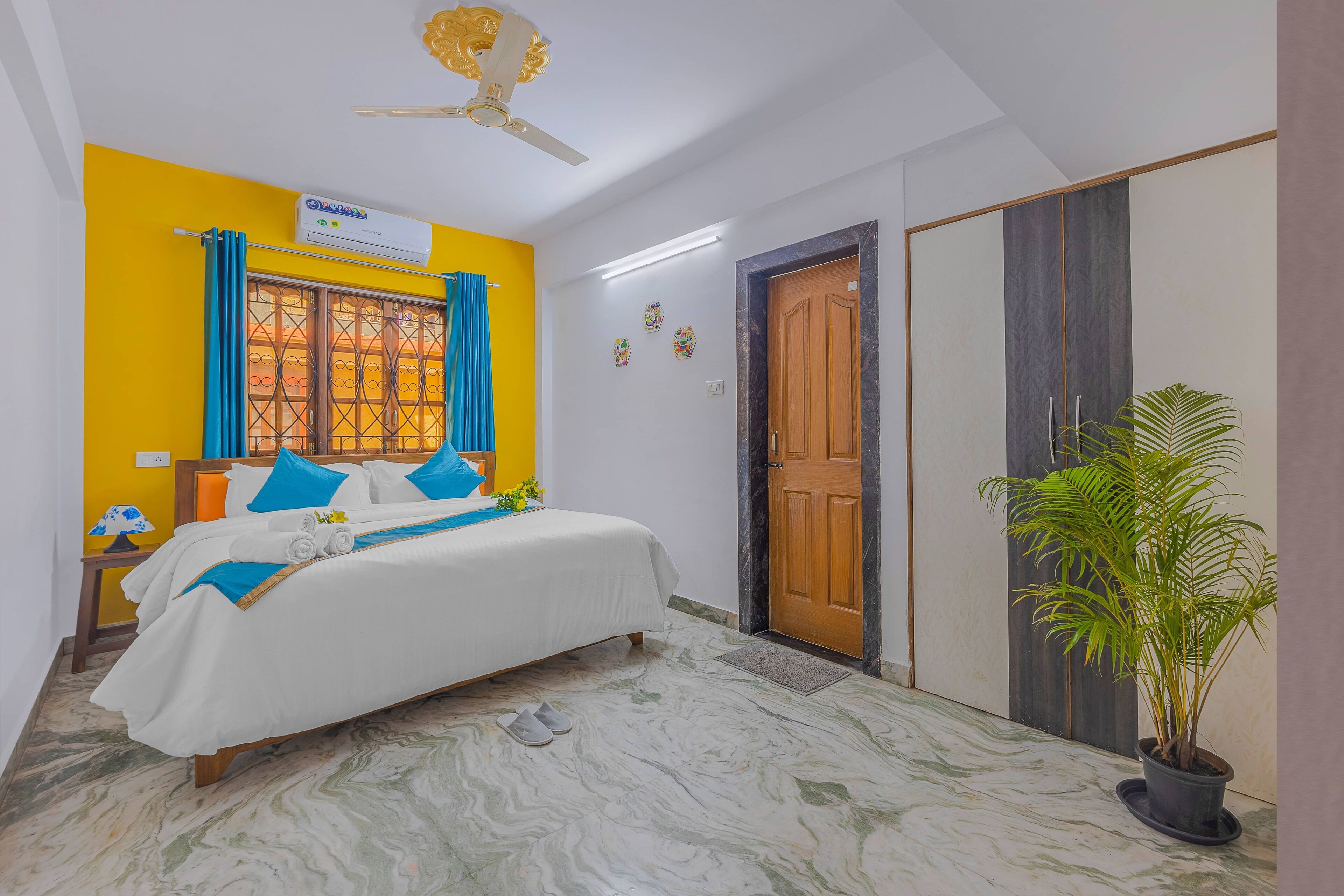 A bedroom with a bed, a ceiling fan, an air conditioning unit, a window with curtains, a door, and a potted plant.
