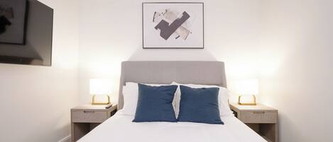 Un lit bien fait, avec des draps blancs et deux oreillers bleus, flanqué de deux tables de chevet surmontées de lampes, dans une pièce au design simple et épuré.